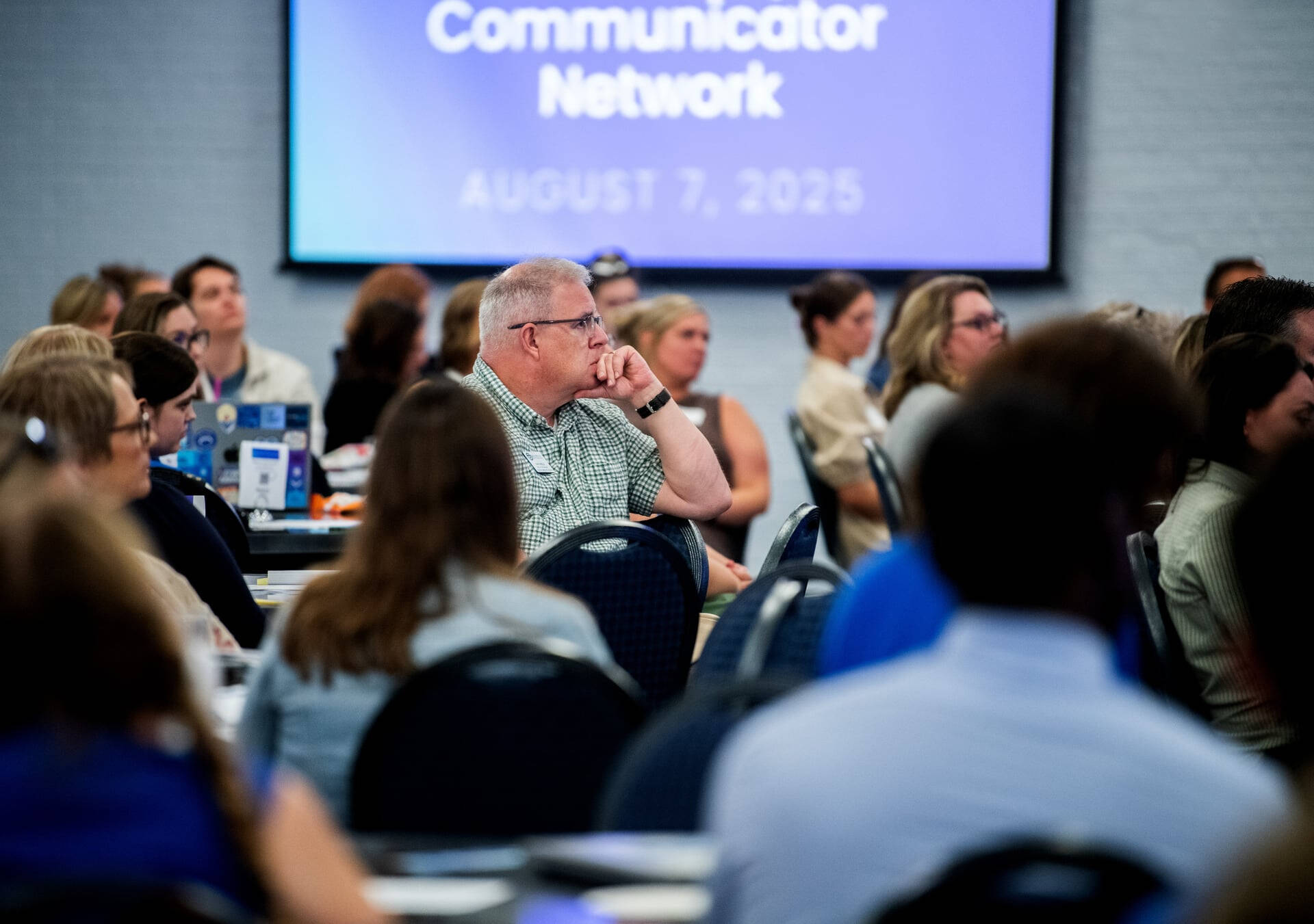 people seated at round tables, projector reads Communicator Network August 7, 2025 in background